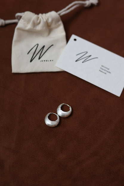 The Minimalist Basic Hoop - The Drop — silver-tone rounded hoops on wood beside a pouch