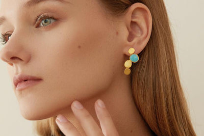 The Stones-Earrings — gold-tone drop earring with blue accent worn on an ear