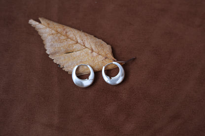 The Minimalist Basic Hoop - The Drop — silver-tone rounded hoops beside a dried leaf